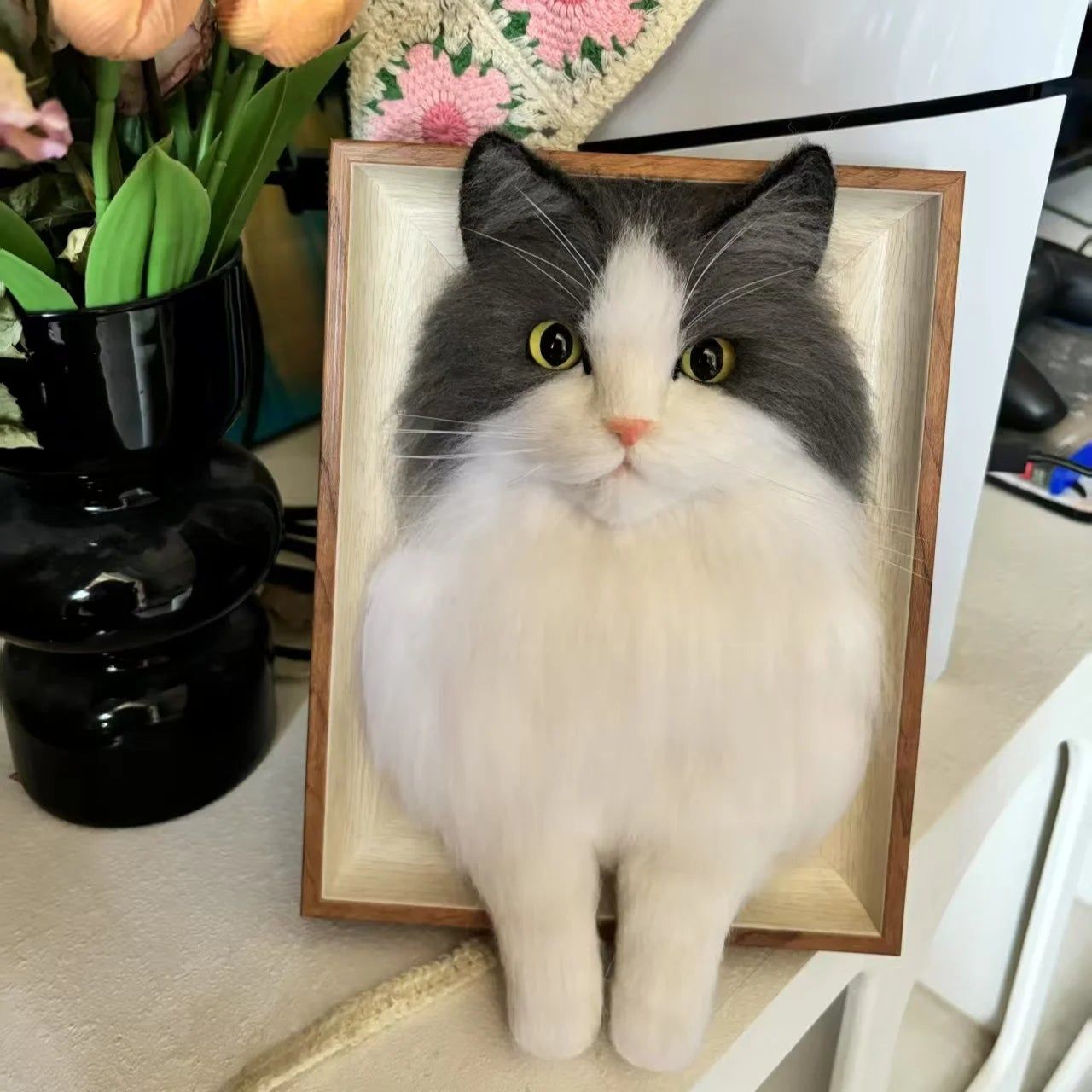 Cat-shaped decorative item in a wooden frame on a table with flowers and a vase in the background.