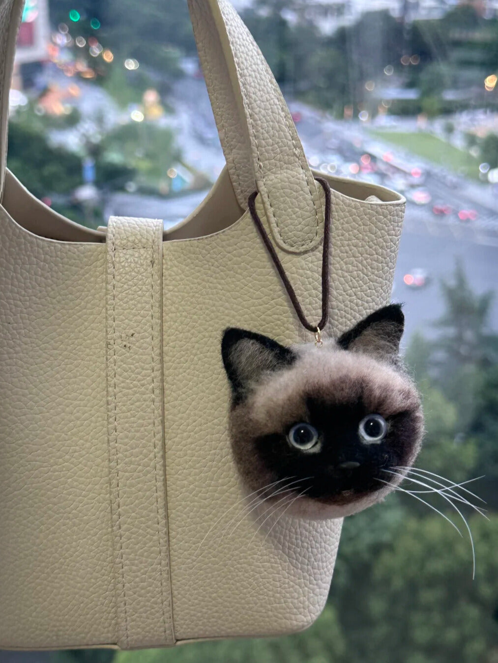Beige handbag with a cat-shaped charm peeking out, blurred cityscape in the background