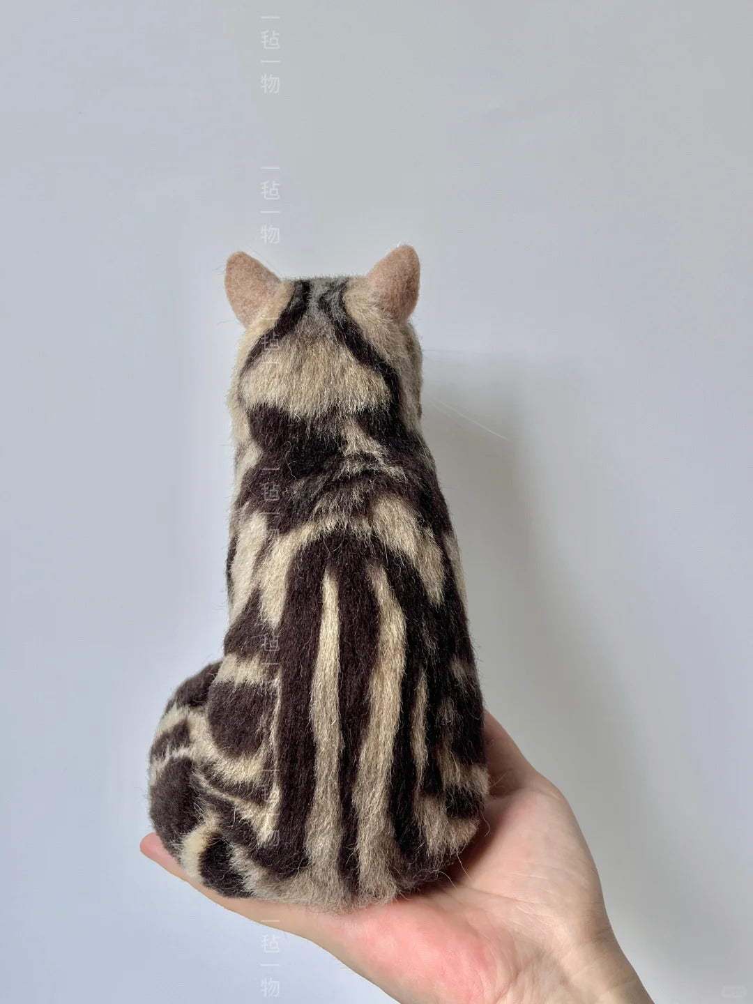 Stuffed animal cat toy with black and brown stripes held in a hand against a light gray background